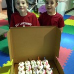 Two boys standing in front of a box full of cupcakes.