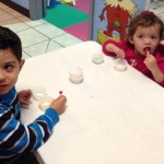 Two children sitting at a table with cups of yogurt.