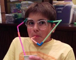 A boy with glasses and drinking straw in his mouth.